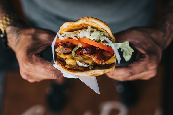 Tattooed hands holding a fully assembled burger wrapped in paper - the end product delivered